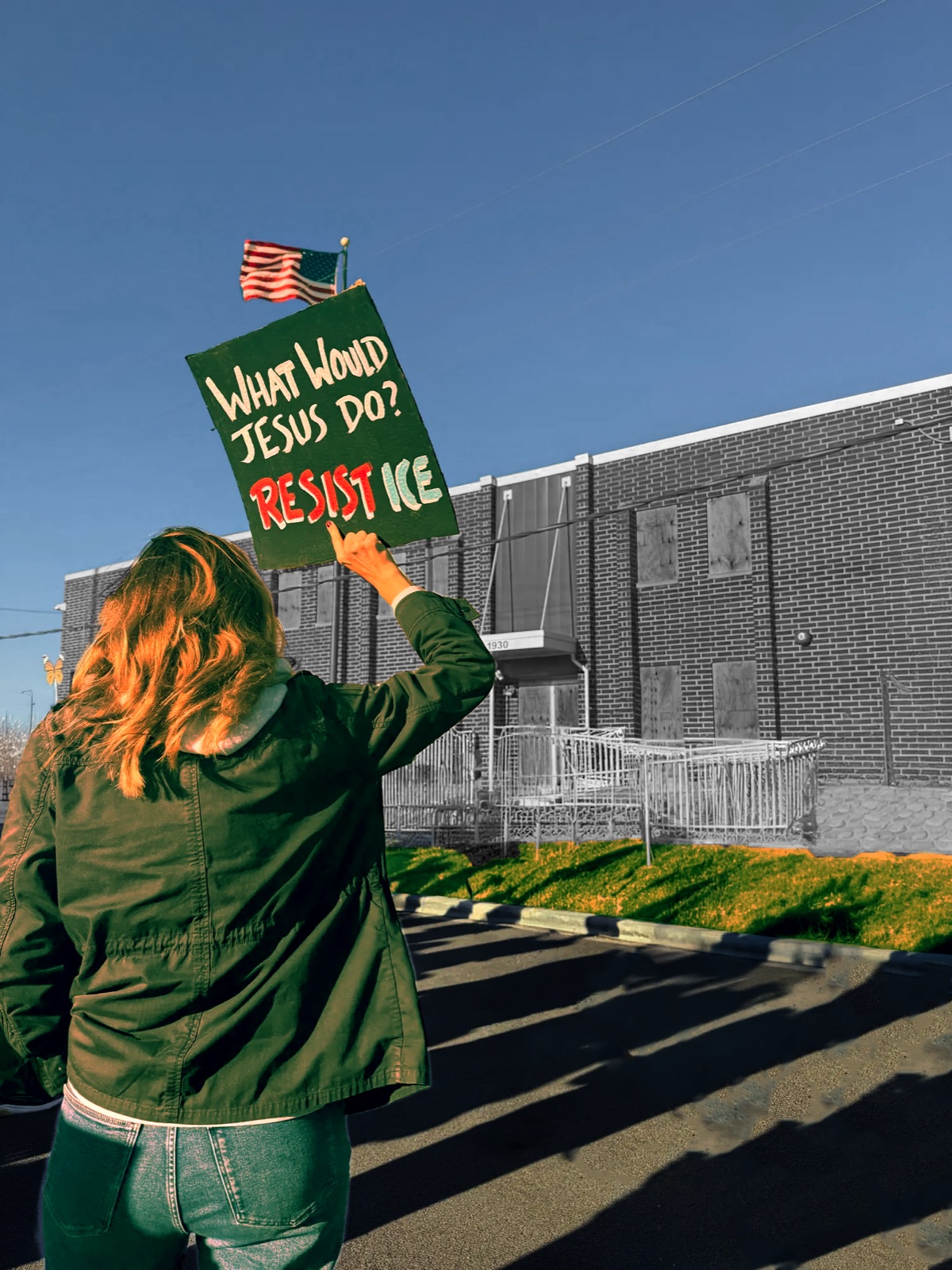 A protester, seen from behind, raises a hand-lettered green sign reading 'WHAT WOULD JESUS DO? RESIST ICE' toward the boarded-up Broadview ICE detention center, an American flag flying overhead against a clear blue sky.